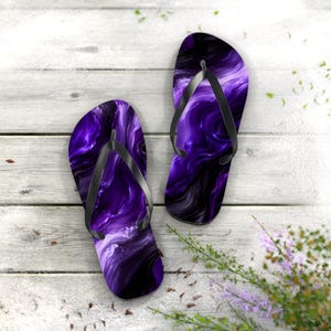 May include: A pair of flip-flops featuring a vibrant purple and black marbled pattern on the footbeds. Each sandal is equipped with a solid black rubber thong strap that secures the foot in place. The design displays swirling liquid-like colors.