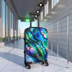 May include: A rolling suitcase featuring a vibrant, iridescent abalone shell pattern in shades of deep blue, teal, and purple. The luggage has a black frame, a retractable handle, and four black spinner wheels, standing upright on a tiled floor.