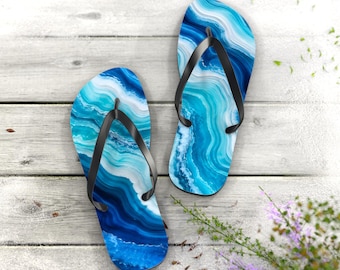 May include: A pair of flip-flops featuring a vibrant blue and white agate-inspired marbled pattern on the footbeds. Each sandal is equipped with a solid black rubber thong strap that secures the foot in place for casual wear.