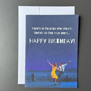 May include: A rectangular greeting card with a dark blue background featuring a couple dancing against a night cityscape. The text reads: Here's to the ones who dream, foolish as they may seem... HAPPY BIRTHDAY! A white envelope is positioned behind the card.