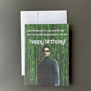 May include: A rectangular greeting card with a white envelope featuring a digital rain background in shades of green. The card displays a figure in a dark coat and sunglasses with the text: take the blue pill it's just another day, take the red pill party like you're the one, happy birthday!