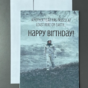 May include: A rectangular greeting card featuring a blue-toned illustration of an astronaut walking across a rocky landscape. The text reads: ANOTHER YEAR HAS PASSED, AT LEAST HERE ON EARTH... HAPPY BIRTHDAY! A plain white envelope is positioned behind the card.