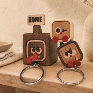 May include: Two wooden keychain holders featuring square characters with dark brown and light wood tones. One holder has a sign that reads HOME. Each character has red circular feet and expressive faces. Two silver metal key rings are attached below.