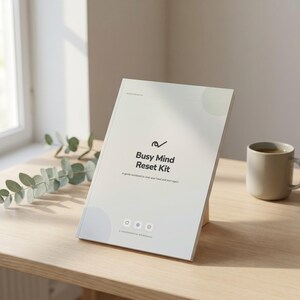 Può includere: Un quaderno di esercizi bianco intitolato Busy Mind Reset Kit con un logo a forma di spunta nera, il sottotitolo A gentle workbook to clear your head and start again e tre piccole icone in basso, esposto su un supporto in legno.