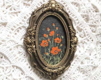 May include: An oval-shaped wall hanging featuring a dark background with a painted illustration of bright orange poppy flowers and green stems, encased in an ornate, antique-style metallic gold frame with intricate scrollwork and shell details.