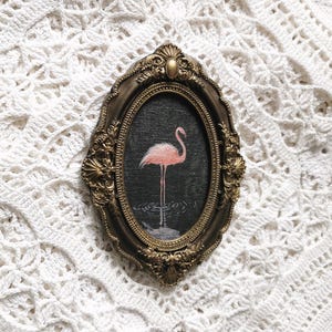 May include: An oval-shaped wall art piece featuring a pink flamingo standing in dark water, encased in an ornate, antique-style gold-toned frame with intricate scrollwork and shell-like details along the edges of the decorative border.
