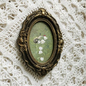 May include: An oval-shaped wall art piece featuring an embroidered snail and white lily of the valley flowers on a green background, encased in an ornate, dark gold-toned frame with intricate scrollwork and a decorative top crest detail.