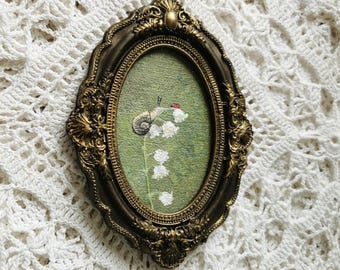 May include: An oval-shaped wall art piece featuring an embroidered snail and white lily of the valley flowers on a green background, encased in an ornate, dark gold-toned frame with intricate scrollwork and a decorative top crest detail.
