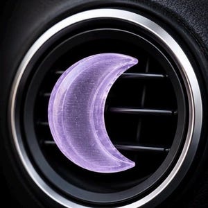 May include: A translucent, light purple crescent moon-shaped object attached to the horizontal slats of a circular black car air vent. The moon has a smooth, glossy surface and is positioned centrally over the dark vent blades.