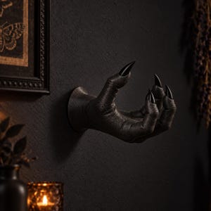 May include: A wall-mounted sculpture of a textured black hand with long, pointed, glossy black fingernails. The hand is positioned in a curved, grasping gesture, extending outward from the wall with a circular base attachment.