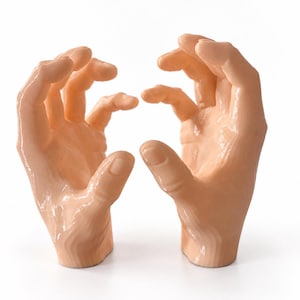 May include: A pair of light peach-colored sculptures shaped like human hands, positioned facing each other with fingers slightly curled and palms open. The surface of the hands has a smooth, glossy finish with visible layered textures.