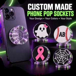 May include: A dark smartphone with a clear, circular phone grip attached, alongside four additional circular phone grips featuring designs of a white ghost with a pink bow, a baseball with the letters AB, a pink ribbon, and a white spiderweb.