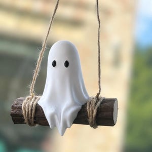 May include: A small white ghost figurine with two black oval eyes, sitting on a dark brown cylindrical log swing. The log is suspended by two strands of natural tan jute twine wrapped around each end, creating a hanging decorative ornament.