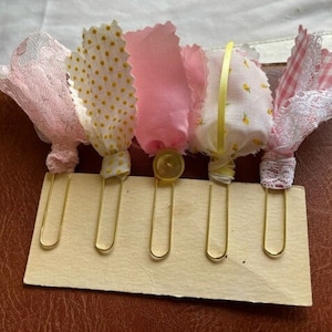 May include: Six gold binder clips with pink and yellow fabric embellishments. The fabric is a mix of polka dots, lace, and gingham.