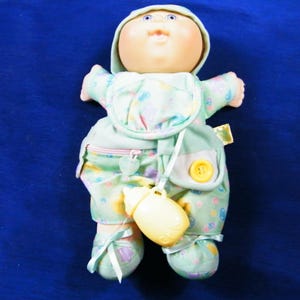 May include: A Cabbage Patch Kids doll with a round face, blue eyes, and a light skin tone. The doll wears a mint green hooded outfit featuring a pastel pattern, a yellow button, a small zippered pocket, and matching booties with ribbons.