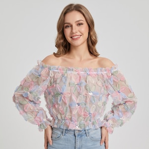 Puede incluir: Una blusa de manga larga con hombros descubiertos hecha de tela transparente con un patrón floral multicolor de pétalos en tonos pastel rosa, azul y verde. Cada flor tiene un pequeño centro de lentejuelas plateadas, cuello y puños elásticos.