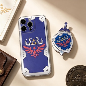 May include: A clear smartphone case featuring a blue shield design with a red bird emblem and a gold triforce symbol, accompanied by a matching circular phone grip shaped like a shield with a sword, both inspired by the Legend of Zelda series.