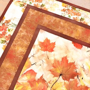 May include: A quilted fabric placemat featuring a central panel with printed orange and red maple leaves on a cream background, framed by a solid dark brown border and an outer section with a delicate orange floral pattern on white fabric.
