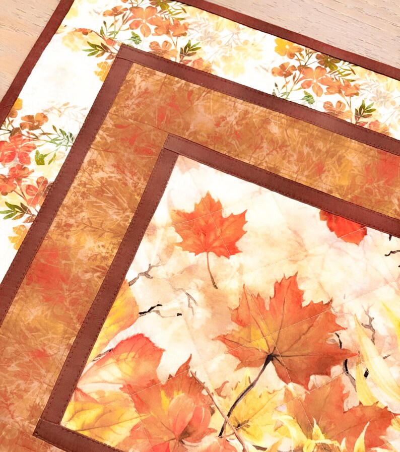 May include: A quilted fabric placemat featuring a central panel with printed orange and red maple leaves on a cream background, framed by a solid dark brown border and an outer section with a delicate orange floral pattern on white fabric.