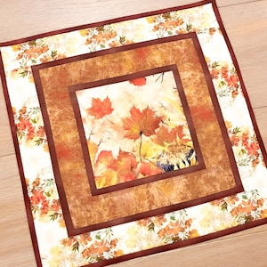 May include: A square fabric table mat featuring a central panel of autumn maple leaves in shades of orange, red, and yellow, surrounded by a textured brown border and an outer frame of white fabric decorated with orange and green floral patterns.