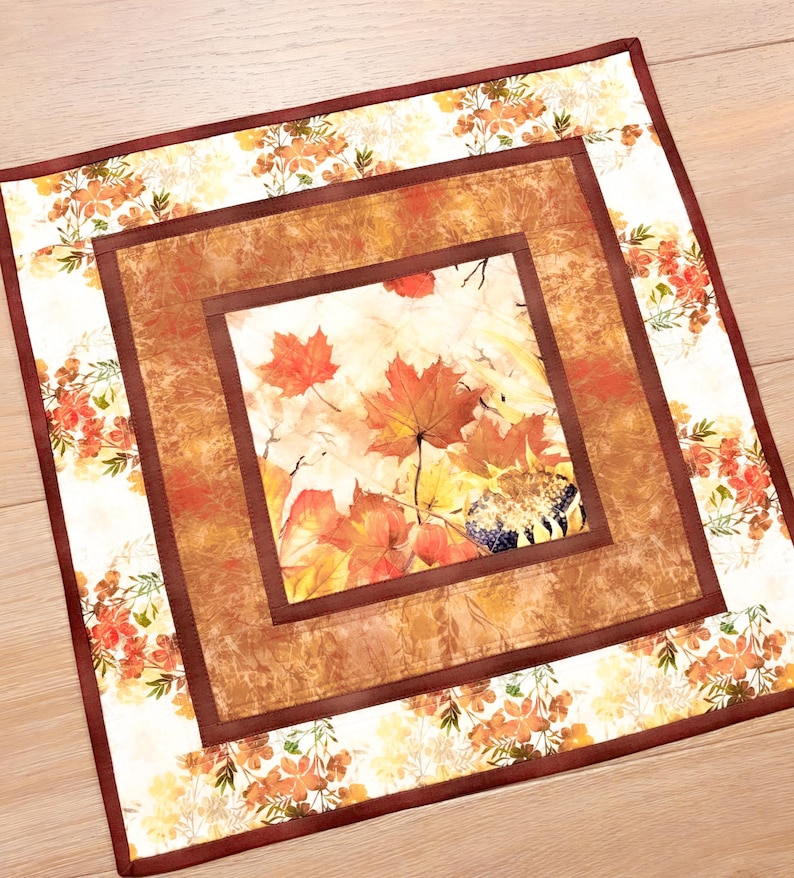 May include: A square fabric table mat featuring a central panel of autumn maple leaves in shades of orange, red, and yellow, surrounded by a textured brown border and an outer frame of white fabric decorated with orange and green floral patterns.