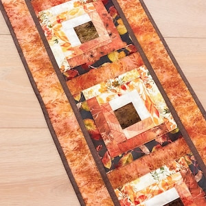 May include: A rectangular fabric table runner featuring a log cabin quilt pattern. The design incorporates various shades of orange, red, yellow, and brown with leaf and foliage prints. The runner is finished with a solid dark brown fabric border.