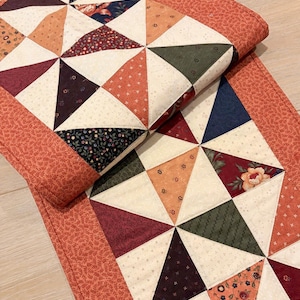 May include: A rectangular quilted table runner featuring a geometric pinwheel pattern composed of triangular fabric patches in cream, orange, dark green, navy blue, and burgundy. The runner is bordered by a solid orange fabric with a small leaf print.