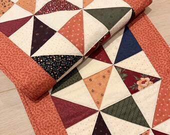 May include: A rectangular quilted table runner featuring a geometric pinwheel pattern composed of triangular fabric patches in cream, orange, dark green, navy blue, and burgundy. The runner is bordered by a solid orange fabric with a small leaf print.