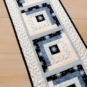 May include: A rectangular quilted table runner featuring four square log cabin blocks in shades of navy blue, light blue, and white. The fabric displays snowflake patterns and delicate floral vines, finished with a solid black border.