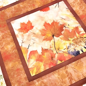 May include: A square quilted fabric mat featuring a central panel with a printed autumn theme of orange and red maple leaves, yellow sunflowers, and dark berries. The center is framed by a solid dark brown border and a wider outer border.