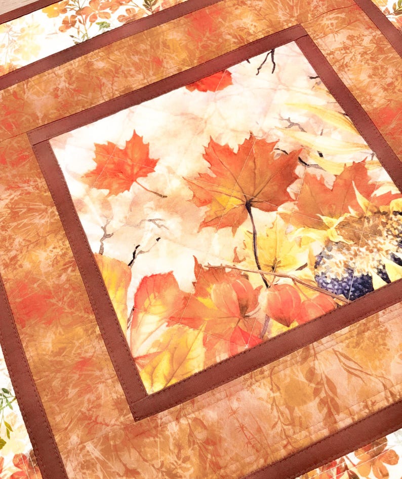May include: A square quilted fabric mat featuring a central panel with a printed autumn theme of orange and red maple leaves, yellow sunflowers, and dark berries. The center is framed by a solid dark brown border and a wider outer border.