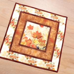 May include: A square fabric table mat featuring a central panel of orange and red maple leaves and a sunflower. The mat has a textured brown border and an outer frame decorated with a pattern of small orange flowers and green foliage.