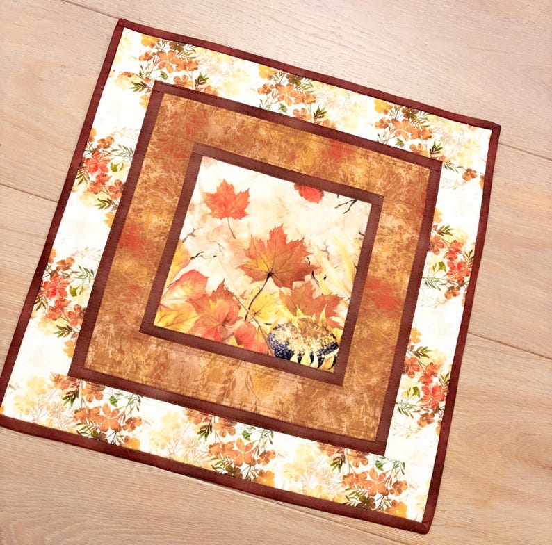 May include: A square fabric table mat featuring a central panel of orange and red maple leaves and a sunflower. The mat has a textured brown border and an outer frame decorated with a pattern of small orange flowers and green foliage.