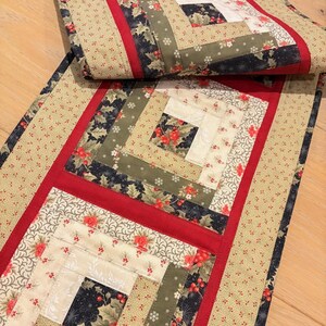 May include: A rectangular table runner featuring a log cabin quilt pattern. The design incorporates fabric squares in shades of cream, olive green, and dark navy, accented with red berries and floral motifs, separated by solid red fabric borders.