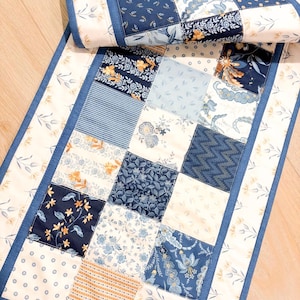 May include: A rectangular table runner featuring a patchwork design of various blue, white, and yellow floral, geometric, and polka dot fabric squares. The runner is bordered by solid blue fabric strips and a cream-colored floral print fabric edge.