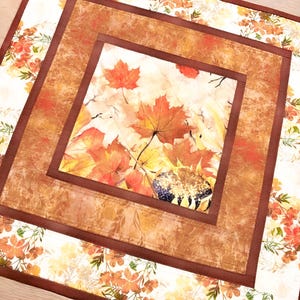 May include: A square fabric table mat featuring a central panel of autumn maple leaves and a sunflower head. The design is framed by a solid brown border and an outer border of white fabric printed with small orange and yellow fall foliage patterns.
