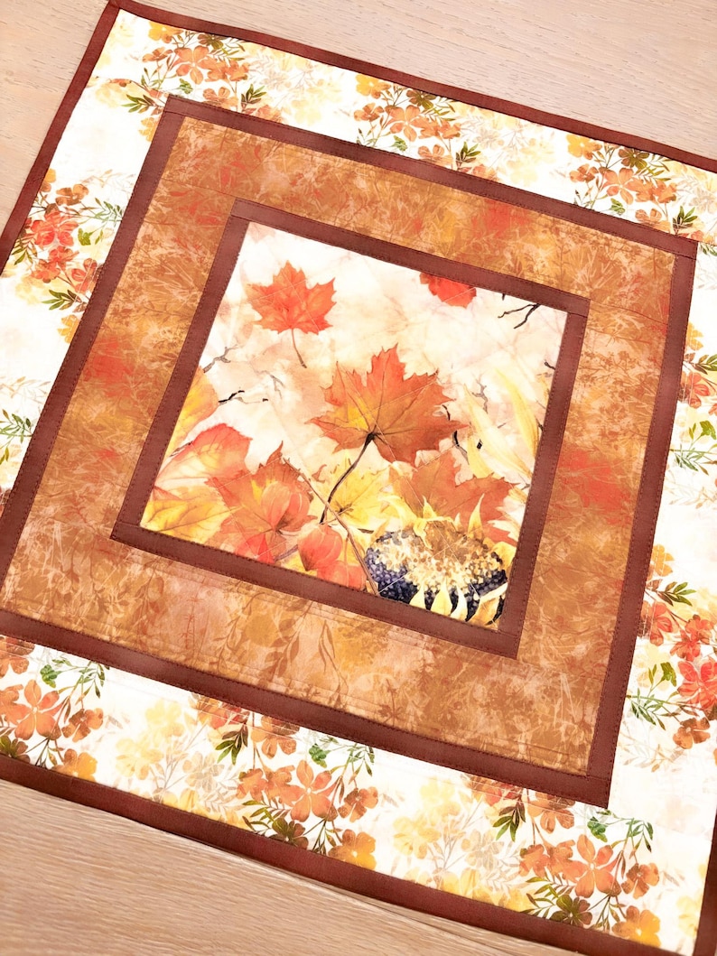 May include: A square fabric table mat featuring a central panel of autumn maple leaves and a sunflower head. The design is framed by a solid brown border and an outer border of white fabric printed with small orange and yellow fall foliage patterns.
