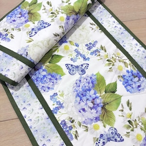 May include: A quilted table runner featuring a floral design with blue hydrangeas, white flowers, and butterflies on a white background. The edges are trimmed with a dark green border. The runner is folded over, showing the pattern.