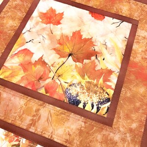May include: A square fabric table mat featuring a central panel with a printed autumn motif of orange and red maple leaves and a sunflower. The central panel is framed by a solid dark brown border and an outer border with a leaf pattern.