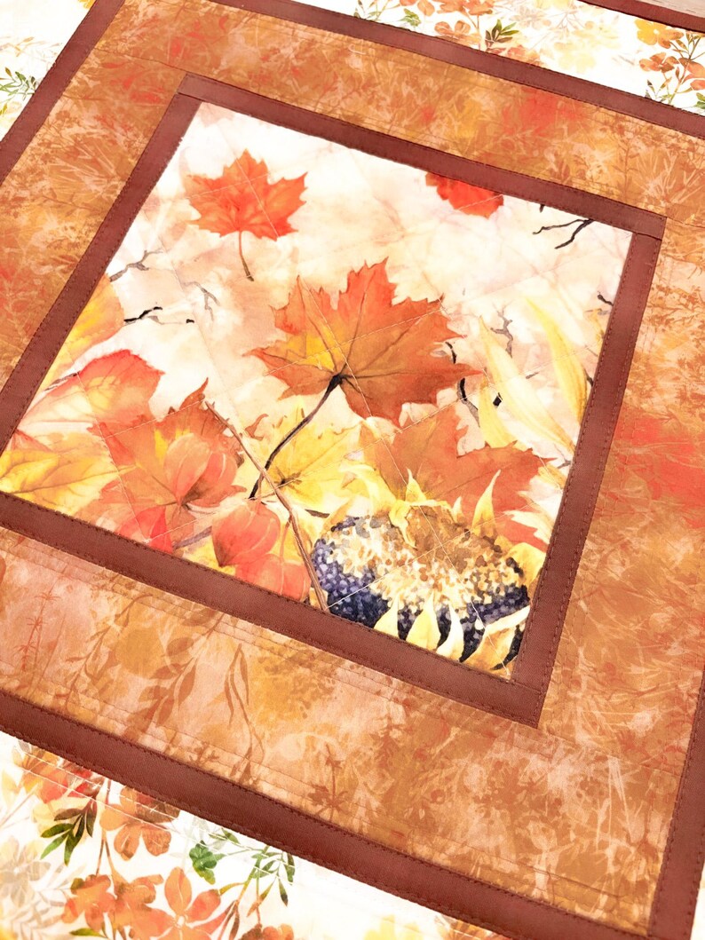 May include: A square fabric table mat featuring a central panel with a printed autumn motif of orange and red maple leaves and a sunflower. The central panel is framed by a solid dark brown border and an outer border with a leaf pattern.
