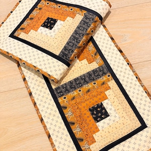 May include: A rectangular fabric table runner featuring a log cabin quilt pattern. The design incorporates strips of fabric in shades of mustard yellow, cream, and black with floral and leaf prints, bordered by a cream fabric with a small black cross pattern.