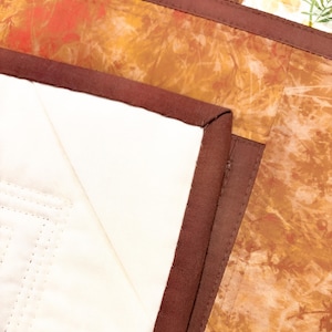 May include: A quilted textile piece featuring a solid cream-colored section and a contrasting section with an orange and brown mottled pattern. The two sections are joined by a dark brown fabric binding with visible straight-line stitching.