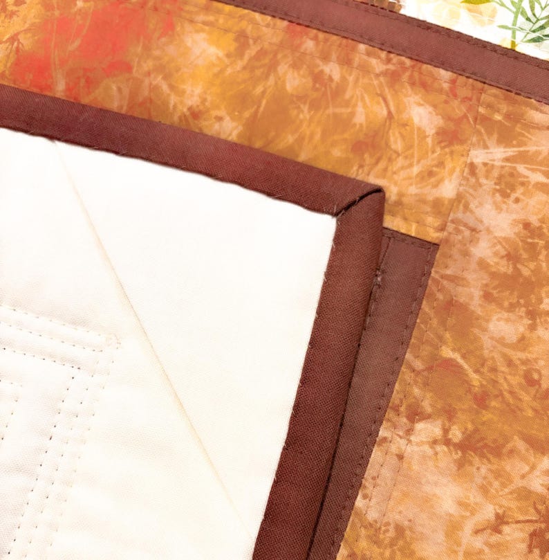May include: A quilted textile piece featuring a solid cream-colored section and a contrasting section with an orange and brown mottled pattern. The two sections are joined by a dark brown fabric binding with visible straight-line stitching.