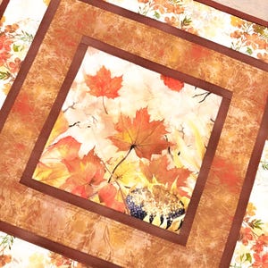 May include: A square fabric table mat featuring a central panel with a print of orange and red maple leaves and a sunflower. The center is framed by a solid brown border, surrounded by an outer layer with a white background and floral print.