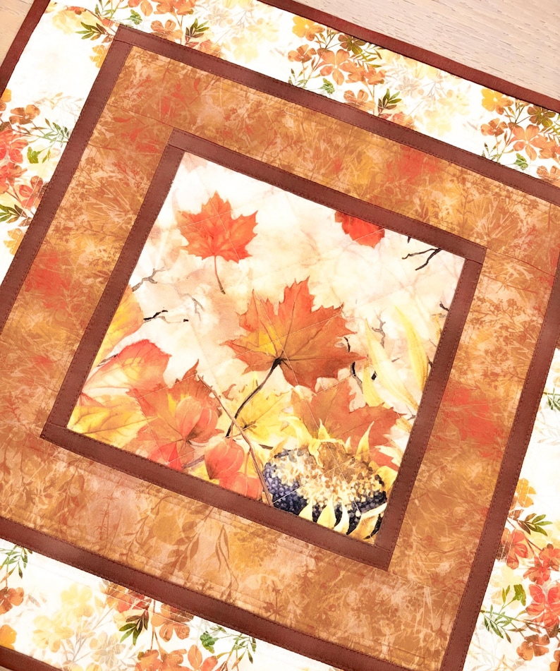 May include: A square fabric table mat featuring a central panel with a print of orange and red maple leaves and a sunflower. The center is framed by a solid brown border, surrounded by an outer layer with a white background and floral print.