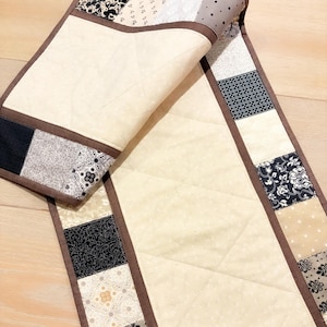 May include: A quilted table runner with a cream-colored center and a brown border. The border features a patchwork design with black, white, and tan patterned squares. The runner is folded over, revealing the quilted underside.
