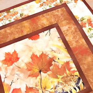 May include: A square quilted fabric placemat featuring a central panel with orange and red maple leaves and sunflowers, framed by a solid dark brown border. The fabric has a visible stitched grid pattern and a light cream background throughout.