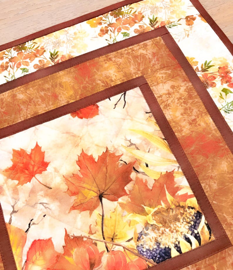 May include: A square quilted fabric placemat featuring a central panel with orange and red maple leaves and sunflowers, framed by a solid dark brown border. The fabric has a visible stitched grid pattern and a light cream background throughout.