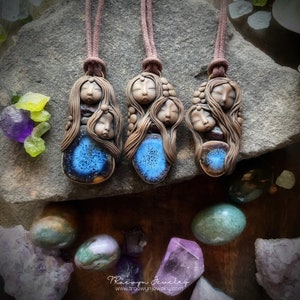 Puede incluir: Tres colgantes de arcilla con detalles azules y marrones, cada uno con tres caras dormidas con cabello largo. Los colgantes cuelgan de cordones marrones y se muestran sobre una superficie de roca con varias piedras preciosas.