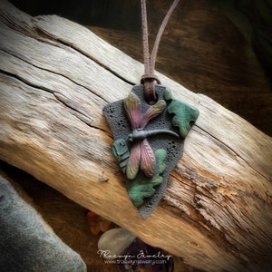May include: A dark grey, textured, arrowhead-shaped pendant with a red and black dragonfly and green leaves. The pendant is on a brown leather cord.